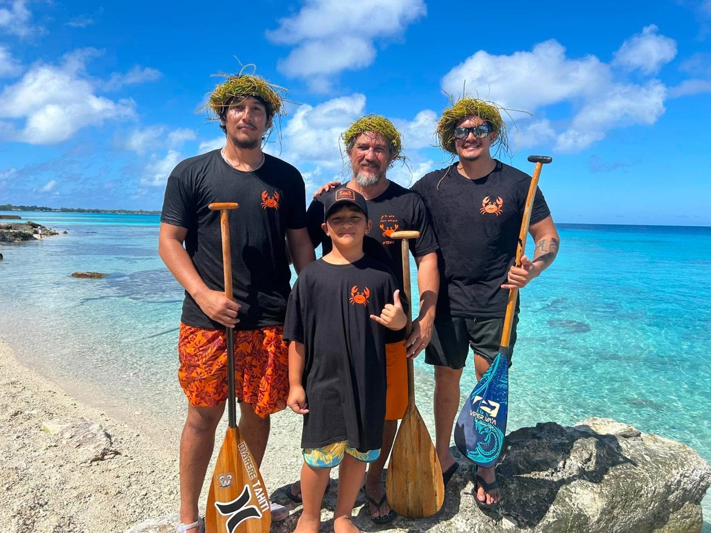 Trois rameurs et un enfant en tee-shirts au crabe, pagaies à la main, au bord du lagon