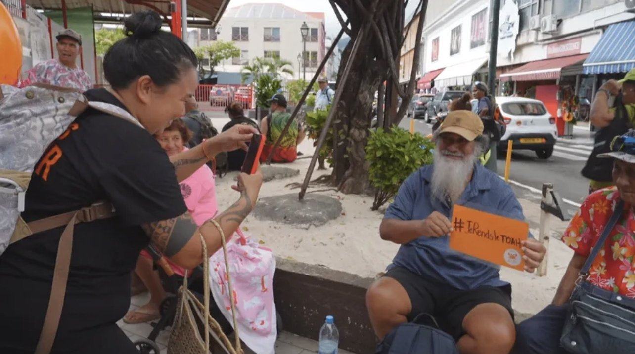 Une maman de l'association photographie un passant tenant une pancarte de sensibilisation au marché de Papeete