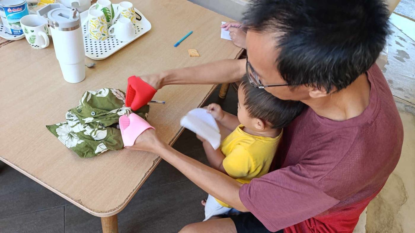 Un papa, son enfant sur les genoux, essaie de boutonner une chemise avec des gants